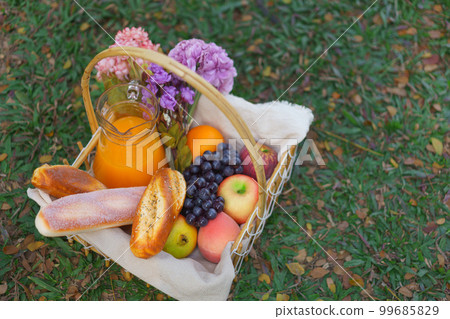 Wicker picnic basket of bakery fruits and orange juice with flowers on grass in the park 99685829