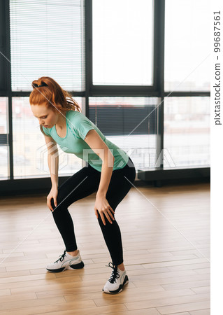 Vertical shot of athletic young woman doing squats exercising and stop training as feeling sharp knee pain during fitness workout at home by window. Female athlete sportswoman touching leg in injury. Vertical shot of athletic young woman doing squats exercising and stop training as feeling sharp knee pain during fitness workout at home by window. Female athlete sportswoman touching leg in injury. 99687561