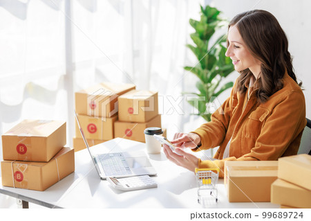 Concept of parcel delivery and selling online,Female seller or retailer using smartphone to check parcel box before delivery and using laptop checking day's sales on the online store Concept of parcel delivery and selling online,Female seller or retailer using smartphone to check parcel box before delivery and using laptop checking day's sales on the online store 99689724