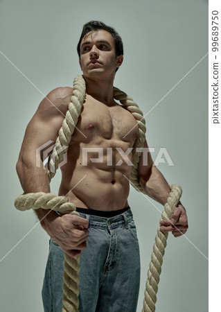 Handsome young man with strong muscular body shape posing in jeans with rope over grey studio background. Concept of men's health, beauty, sport Handsome young man with strong muscular body shape posing in jeans with rope over grey studio background. Concept of men's health, beauty, sport 99689750