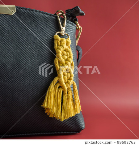 Close-up view of the handmade Macrame key chain hanging on the black hand bag isolated on the red background. Close-up view of the handmade Macrame key chain hanging on the black hand bag isolated on the red background. 99692762