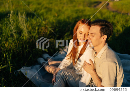Loving young couple hugging with closed eyes enjoying their time together sitting in field on background of warm sunlight in summer evening during sunset. Happy man and woman having romantic date. 99694253
