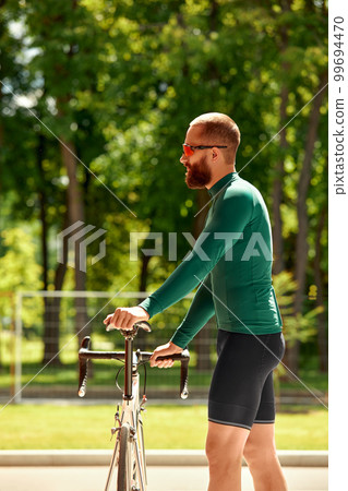 A professional cyclist in sportswear with glasses is preparing for a morning bike ride in a green city park. The concept of an active and healthy lifestyle. 99694470