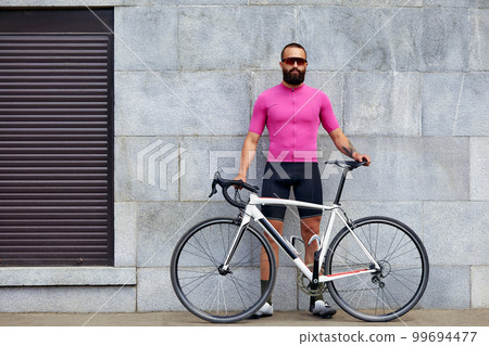 A bearded man cyclist in glasses with a bicycle on the background of a gray wall in the urban landscape, a daily bike ride. A bearded man cyclist in glasses with a bicycle on the background of a gray wall in the urban landscape, a daily bike ride. 99694477