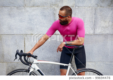 A bearded man cyclist in glasses with a bicycle on the background of a gray wall in the urban landscape, a daily bike ride. 99694478