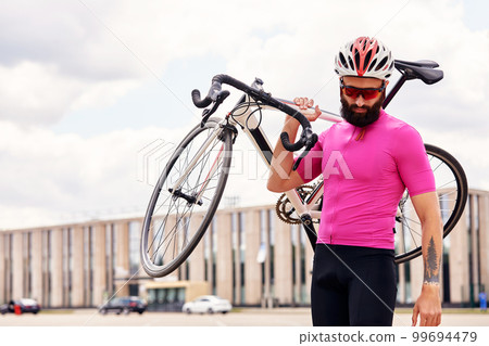 Bearded male cyclist in glasses and a helmet looks at the camera with a bicycle in his hand against the blue sky, lifestyle, cardio workout Bearded male cyclist in glasses and a helmet looks at the camera with a bicycle in his hand against the blue sky, lifestyle, cardio workout 99694479