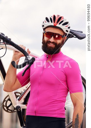 Bearded male cyclist in glasses and a helmet looks at the camera with a bicycle in his hand against the blue sky, lifestyle, cardio workout Bearded male cyclist in glasses and a helmet looks at the camera with a bicycle in his hand against the blue sky, lifestyle, cardio workout 99694480
