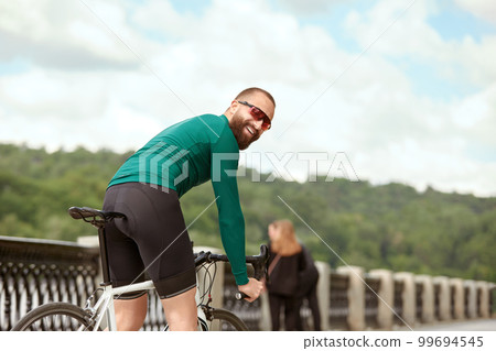 A professional cyclist in sportswear with glasses is preparing for a morning bike ride in a green city park. The concept of an active and healthy lifestyle. 99694545