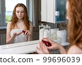 Red-haired girl applies cream to her face looking at herself in the bathroom mirror after shower 99696039