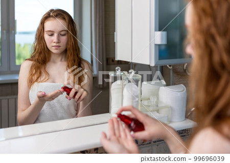 Red-haired girl applies cream to her face looking at herself in the bathroom mirror after shower 99696039