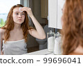 Red-haired teenage girl wipes her face with lotion after shower looking at herself in the bathroom mirror 99696041