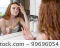 Red-haired girl applies cream to her face looking at herself in the bathroom mirror after shower 99696054