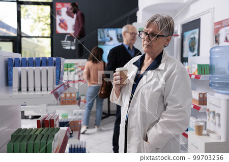 Elderly drugstore worker holding cup of coffee while looking at pharmacy shelves full with vitamins and pills, supplements. Diverse clients doing healthcare shopping session buying medication Elderly drugstore worker holding cup of coffee while looking at pharmacy shelves full with vitamins and pills, supplements. Diverse clients doing healthcare shopping session buying medication 99703256