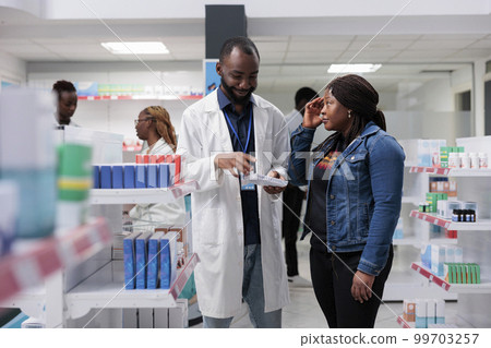 Pharmacy customer suffering from headache, buying painkiller, pharmacy consultant recommending buyer pills. African american woman chatting with pharmacist in drugstore, all black team Pharmacy customer suffering from headache, buying painkiller, pharmacy consultant recommending buyer pills. African american woman chatting with pharmacist in drugstore, all black team 99703257