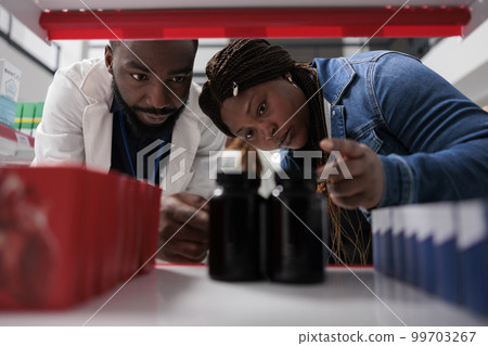 Pharmacist and customer choosing medication on drugstore shelf, close up selective focus. African american woman taking pills packages, pharmaceutical service, medicaments selling Pharmacist and customer choosing medication on drugstore shelf, close up selective focus. African american woman taking pills packages, pharmaceutical service, medicaments selling 99703267