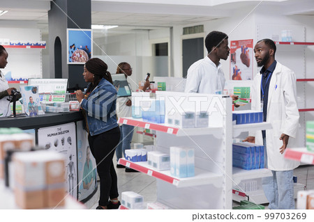 Man choosing vitamins in drugstore, woman buying prescription treatment on checkout counter. African american customers purchasing medicaments in pharmacy store, pharmacist recommending pills Man choosing vitamins in drugstore, woman buying prescription treatment on checkout counter. African american customers purchasing medicaments in pharmacy store, pharmacist recommending pills 99703269