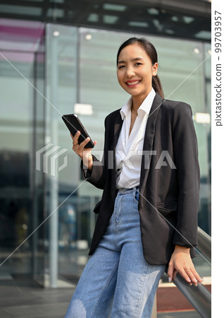 Successful Asian businesswoman holding her smartphone, standing outside of the company building. 99703957