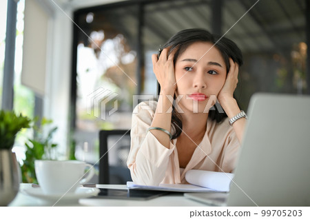 Stressed and thoughtful millennial Asian businesswoman sits at her office desk, pondering 99705203