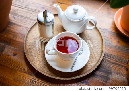 Cup with red fruit tea, teapot and sugar bottle on round wooden tray on wooden window sill, cafe. Top view, close-up. 99706613