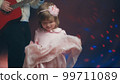 Little girl on stage in pink vintage dress, she dances, her father plays acoustic guitar. Color music is shining. Performance on stage. Early development of children 99711089