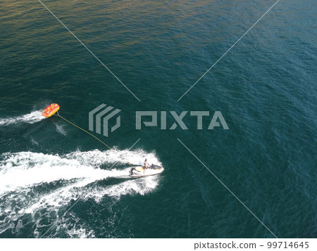 Happy people swim on air mattress behind a high speed water bike. Tourists ride on inflatable watercraft boat. Sea attraction. Water scooter skating people on large Inflatable raft. Aerial copter view Happy people swim on air mattress behind a high speed water bike. Tourists ride on inflatable watercraft boat. Sea attraction. Water scooter skating people on large Inflatable raft. Aerial copter view 99714645