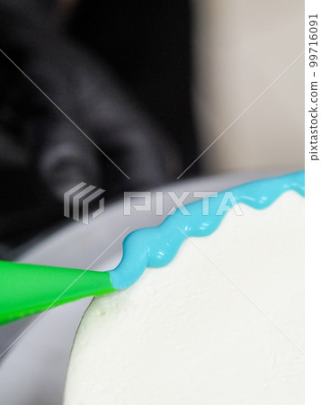 hand pastry chef designer topping a frosted white cake with pastel light blue drip filling bag 99716091