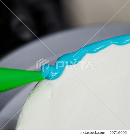 hand pastry chef designer topping a frosted white cake with pastel light blue drip filling bag 99716093