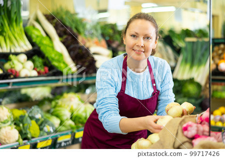 Skillful busy middle-aged female seller in uniform putting fresh onion on shelves in grocery shop Skillful busy middle-aged female seller in uniform putting fresh onion on shelves in grocery shop 99717426