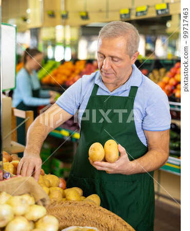 Aged salesman putting potatoes on food stall in greengrocery 99717463