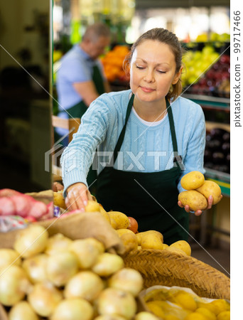 Friendly middle-aged woman market assistant arranging fresh potatoes in grocery supermarket 99717466