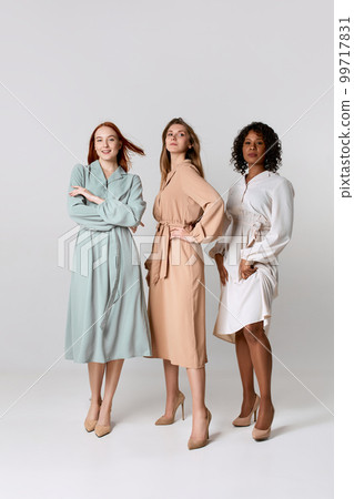 Full-length portrait of diverse beautiful young women in elegant dresses standing together over light background. Concept of spring mood, lightness, beauty, fashion, carefree, ad 99717831