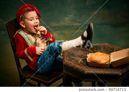 Happy cheerful little boy wearing costume of medieval page boy and prince eating hamburger with french fries over dark green background. Happy cheerful little boy wearing costume of medieval page boy and prince eating hamburger with french fries over dark green background. 99718713