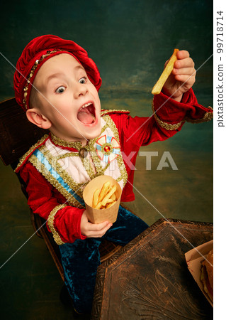 Happy cheerful little boy wearing costume of medieval page boy and prince eating hamburger with french fries over dark green background. 99718714