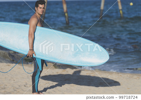 Hispanic surfer boy standing on the beach in neoprene holding his blue surfboard 99718724