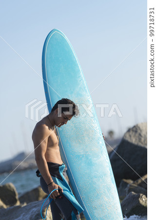 Hispanic surfer boy standing on the beach in neoprene holding his blue surfboard 99718731