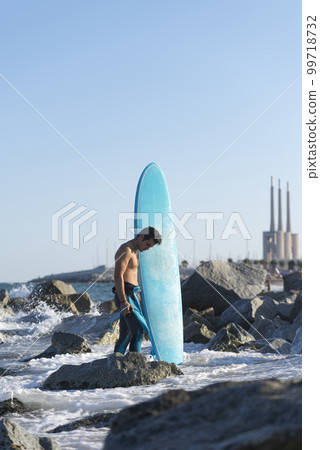 Hispanic surfer boy standing on the beach in neoprene holding his blue surfboard 99718732