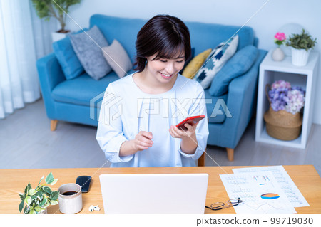 A woman operating a smartphone while using a laptop in the living room A woman operating a smartphone while using a laptop in the living room 99719030