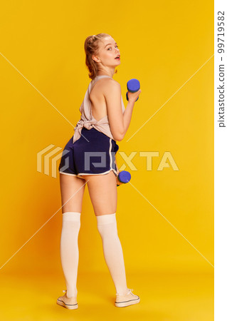 Strength. Full-length image of beautiful slim young girl training with dumbbell over yellow studio background. Concept of sportive lifestyle and emotions 99719582