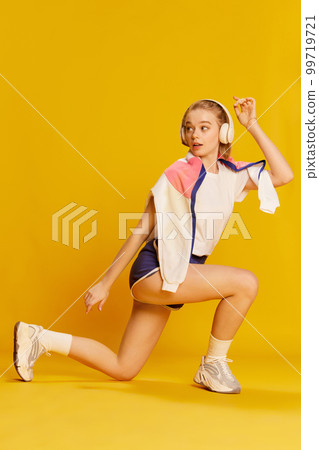 Keeping body fit and healthy. Beautiful young girl in headphones and stylish sportswear training over yellow studio background. Concept of sport lifestyle and emotions Keeping body fit and healthy. Beautiful young girl in headphones and stylish sportswear training over yellow studio background. Concept of sport lifestyle and emotions 99719721