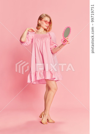Full-length portrait of tender beautiful young girl posing in stylish pink dress, and sunglasses, looking in mirror over pink studio background. Concept of fashion, emotions and beauty Full-length portrait of tender beautiful young girl posing in stylish pink dress, and sunglasses, looking in mirror over pink studio background. Concept of fashion, emotions and beauty 99719751