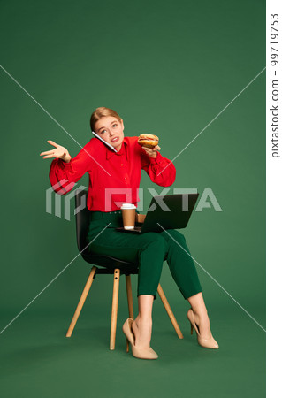 Young beautiful woman, employee talking on phone, working on laptop and eating over green studio background. Many tasks. Concept of business, emotions and lifestyle Young beautiful woman, employee talking on phone, working on laptop and eating over green studio background. Many tasks. Concept of business, emotions and lifestyle 99719753