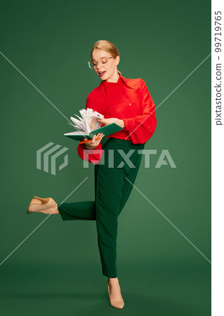 Beautiful young girl, business woman in glasses and red stylish blouse posing over green studio background. Looking through notes. Concept of business, emotions and lifestyle 99719765