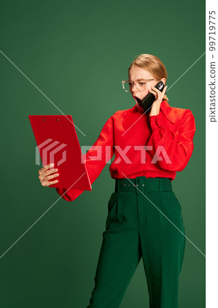 Busy days. Young girl, employee emotionally talking on phone and looking at document over green studio background. Concept of business, emotions and lifestyle Busy days. Young girl, employee emotionally talking on phone and looking at document over green studio background. Concept of business, emotions and lifestyle 99719775