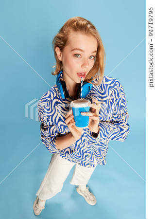 Tender cute young girl in headphones and casual clothes drinking coffee, posing over blue studio background. Delightful look 99719796