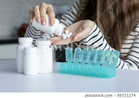 A young woman pours white pills into her hand in front of a pill organizer. Nearby mock-ups of white cans of supplements. A young woman pours white pills into her hand in front of a pill organizer. Nearby mock-ups of white cans of supplements. 99721274