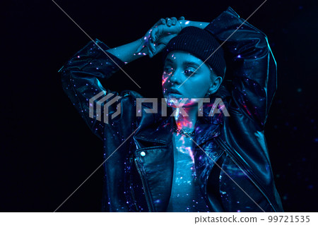 Portrait of young blonde girl with neon space holographic projection on body posing over dark background in blue neon lights. Cyberpunk style Portrait of young blonde girl with neon space holographic projection on body posing over dark background in blue neon lights. Cyberpunk style 99721535