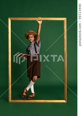 African little boy, child in checkered shirt and suspender shorts standing near huge picture frame over dark green background. Concept of childhood, friendship, retro fashion style 99723490