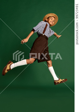 Happy little boy, child in checkered shirt and suspender shorts jumping, having fun over dark green background. Concept of childhood, friendship, retro fashion style Happy little boy, child in checkered shirt and suspender shorts jumping, having fun over dark green background. Concept of childhood, friendship, retro fashion style 99723491
