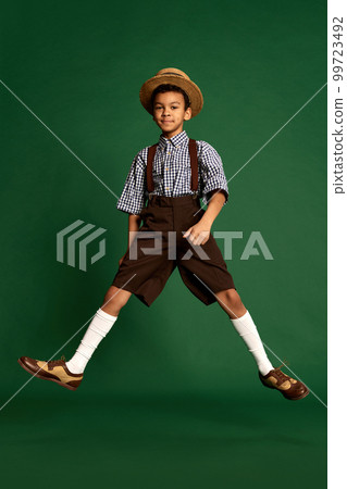 Happy little boy, child in checkered shirt and suspender shorts jumping, having fun over dark green background. Concept of childhood, friendship, retro fashion style 99723492