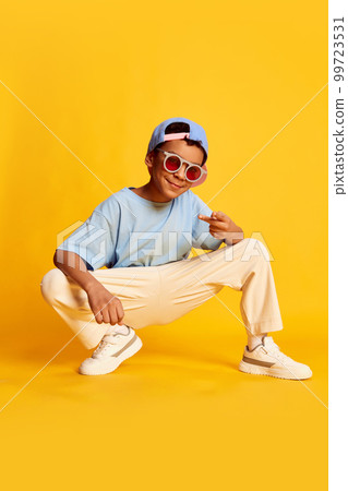 Dynamic portrait of little african boy, hip-hop dancer in stylish street style clothes sitting over bright yellow background. Concept of music, dance happiness Dynamic portrait of little african boy, hip-hop dancer in stylish street style clothes sitting over bright yellow background. Concept of music, dance happiness 99723531